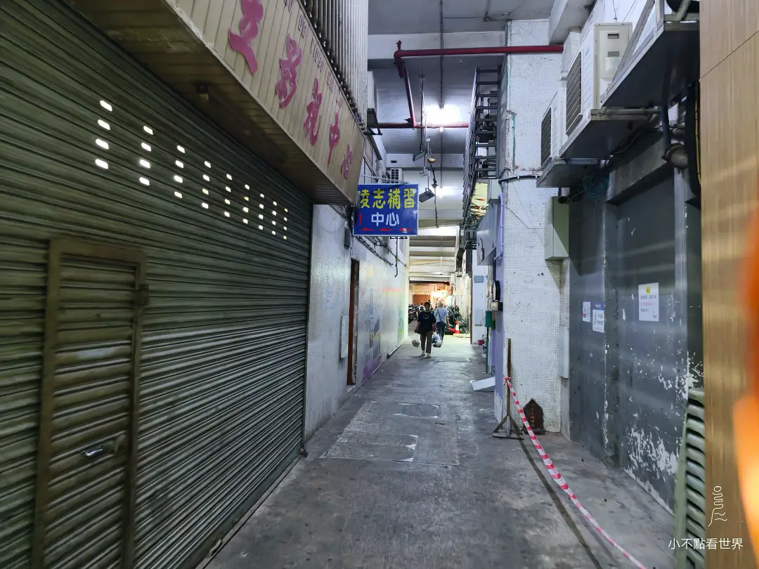澳門一條狹窄的舊式巷弄，兩旁是緊閉的鐵捲門與建築物，遠處可見行人與藍色招牌，呈現當地老街風貌。