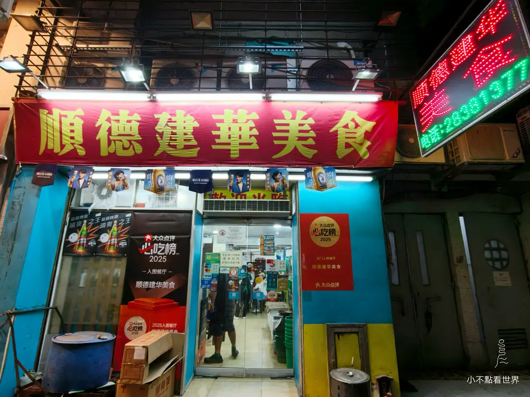 澳門順德建華美食的紅色招牌高掛於店面入口上方，藍色牆面與開著的門口可見店內景象，右側有霓虹燈招牌，並貼有「大眾點評必吃榜2025」的標誌。