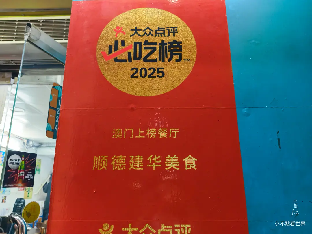一張紅底金字的招牌，上面印有「大眾點評必吃榜2025」的金色圓形標誌，下方文字顯示「澳門上榜餐廳 順德建華美食」。