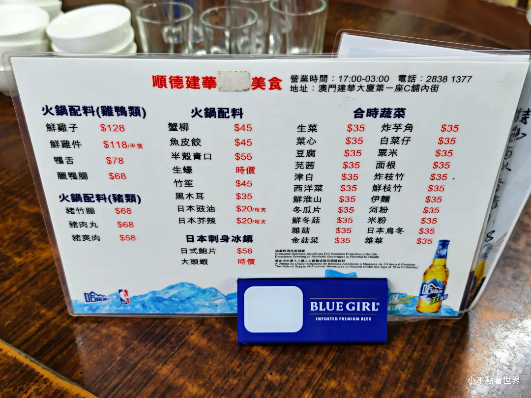 順德建華美食的菜單，列出火鍋配料如鮮雞子、鵝腸、豬肉丸等，以及合時蔬菜如生菜、芥蘭、油麥菜等，並標示價格。前景有藍妹啤酒的杯墊。