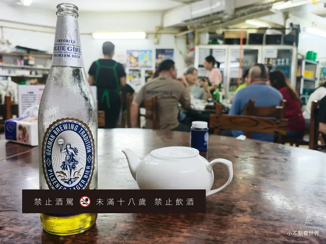 澳門順德建華美食餐廳內，一張木桌上擺放著一瓶冰涼的藍妹啤酒和一個白色茶壺，背景可見模糊的用餐顧客。